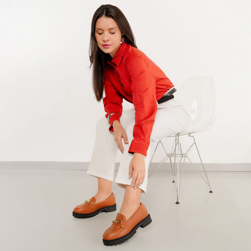 Modelo usando mocassim M Shuz Whisk Napa Soft marrom mostrando ambos os pés com sola preta tratorada e detalhe de corrente dourada.